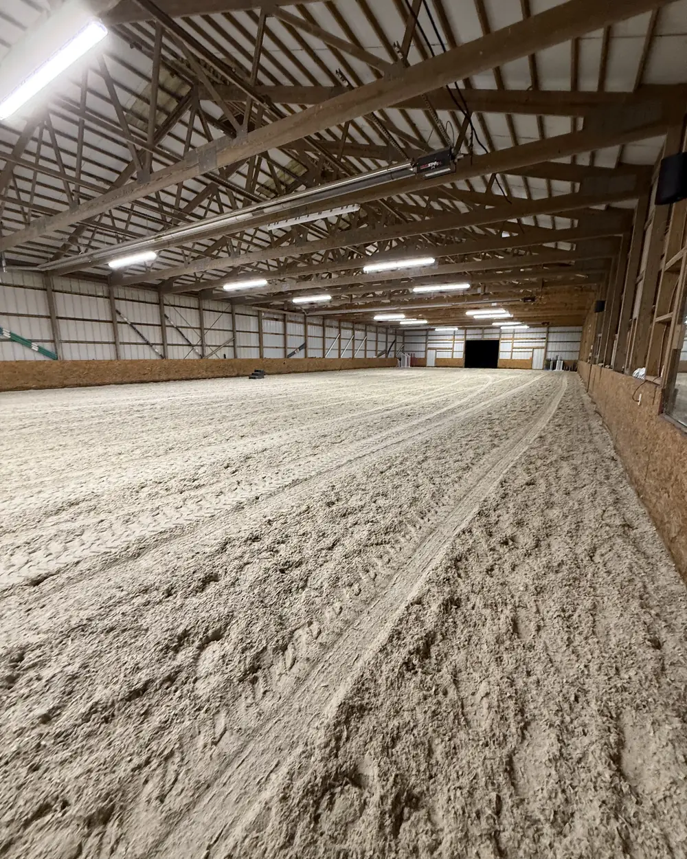The inside of a large steel-framed riding arena, its sand footing graded flat from end to end with machine tracks still running down it
