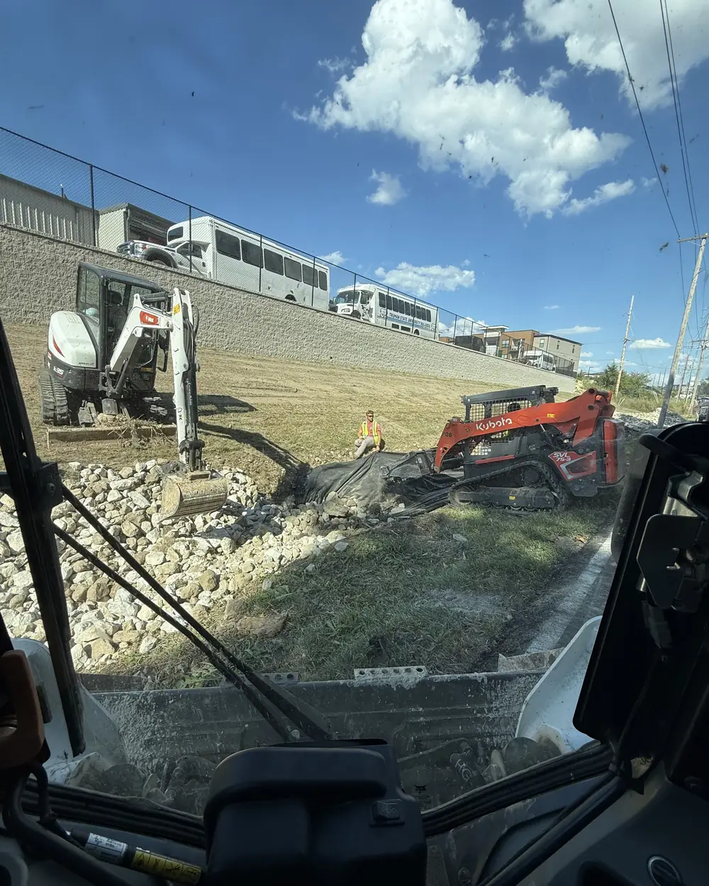 A mini excavator and a tracked skid steer working a rock-lined drainage channel below a retaining wall, with a man in a hi-vis vest on the bank between them