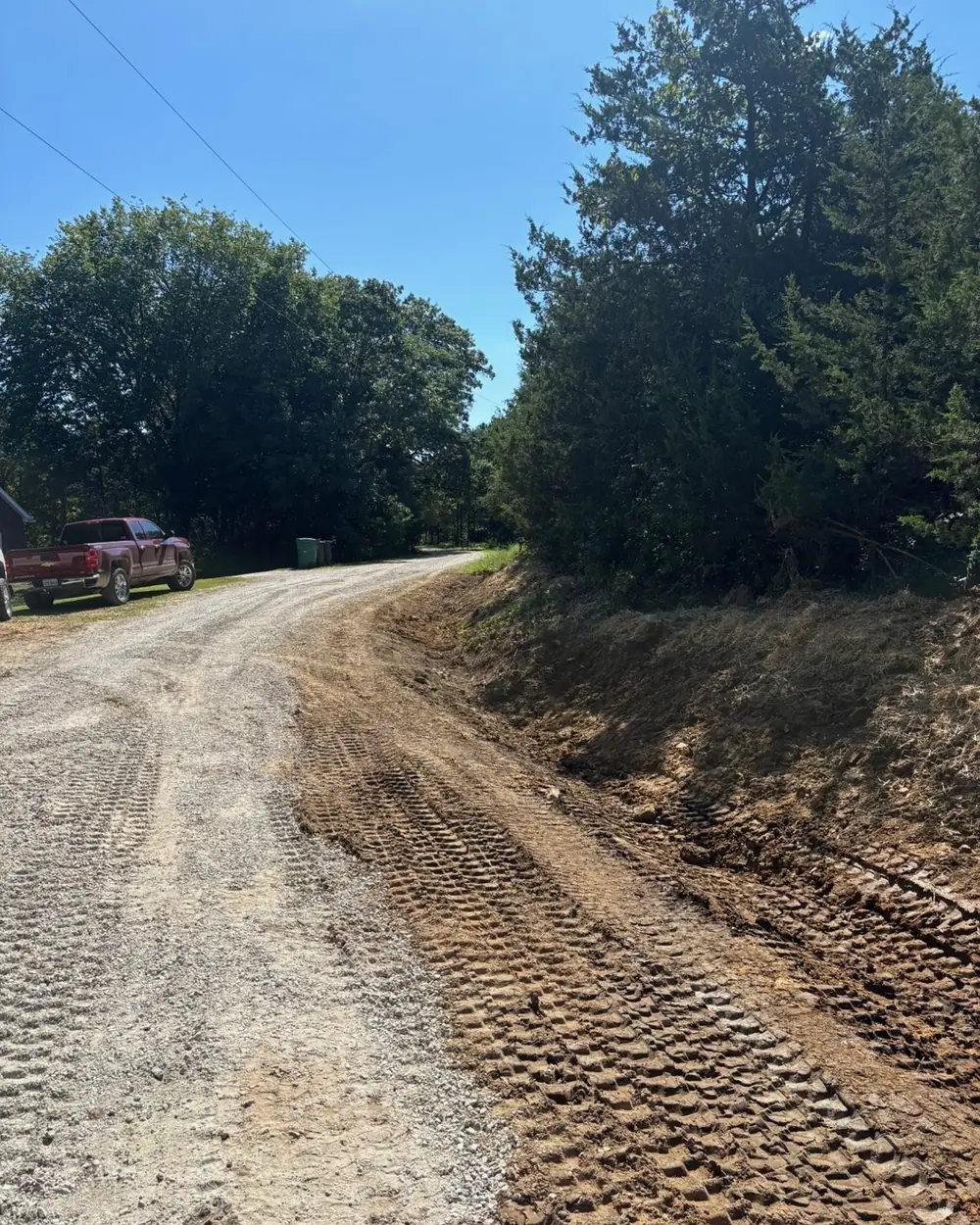 A freshly cut drainage ditch running along the edge of a gravel driveway, the new channel shaped and the spoil graded back